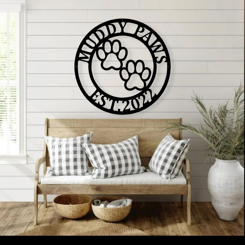 May include: Black metal wall art with the words "Muddy Paws" and "EST. 2022" surrounding two paw prints. The sign is displayed above a wooden bench with checkered pillows and woven baskets.