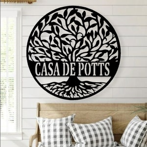 May include: A black metal tree of life wall art with the text "CASA DE POTTS" in a circular design. The tree features detailed leaves and roots. The artwork is mounted on a white shiplap wall above a wooden bench with pillows.