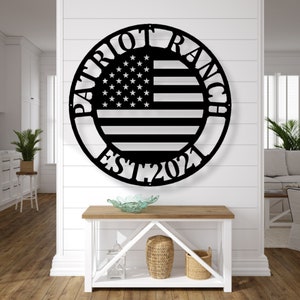 K&ouml;nnte beinhalten: Schwarze Metall-Wandkunst mit dem Design der amerikanischen Flagge in einem kreisf&ouml;rmigen Rahmen. Die Worte "PATRIOT RANCH" sind oben gebogen, mit "EST 2021" unten. Das Kunstwerk ist an einer wei&szlig;en Wand &uuml;ber einem Holztisch angebracht.