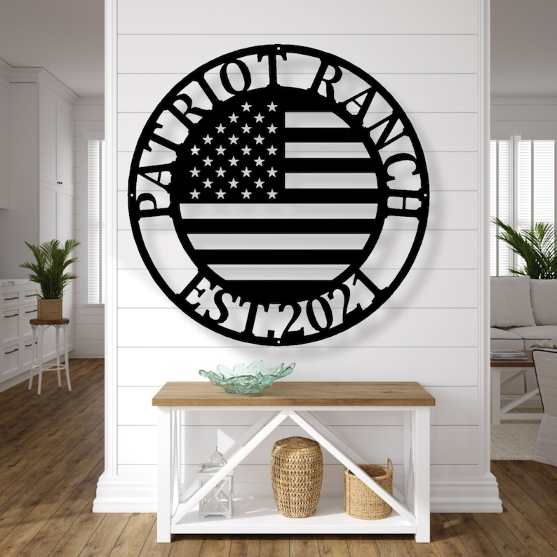 K&ouml;nnte beinhalten: Schwarze Metall-Wandkunst mit dem Design der amerikanischen Flagge in einem kreisf&ouml;rmigen Rahmen. Die Worte "PATRIOT RANCH" sind oben gebogen, mit "EST 2021" unten. Das Kunstwerk ist an einer wei&szlig;en Wand &uuml;ber einem Holztisch angebracht.