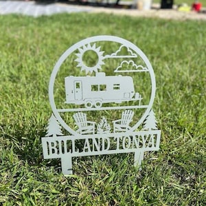 May include: A white metal yard stake featuring a camper, sun, clouds, and two chairs around a campfire. The text "BILL AND TONYA" is displayed on the bottom of the stake. The stake is placed in green grass.