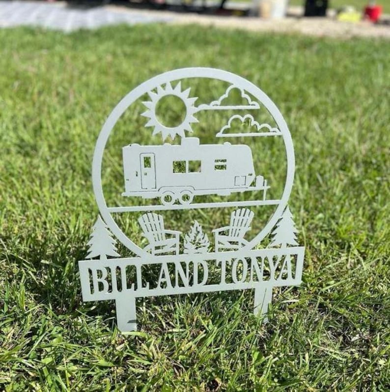 May include: A white metal yard stake featuring a camper, sun, clouds, and two chairs around a campfire. The text "BILL AND TONYA" is displayed on the bottom of the stake. The stake is placed in green grass.
