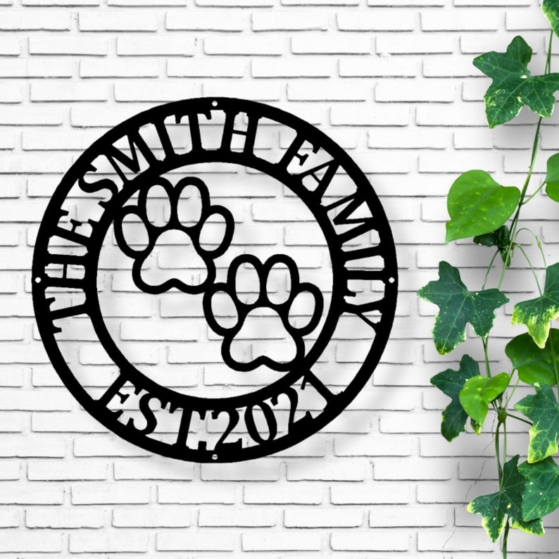 May include: Black metal family sign with the text "THE SMITH FAMILY EST 2021" in a circular design. Two paw prints are in the center. The sign is mounted on a white brick wall.