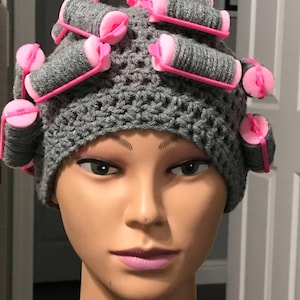 May include: A gray crocheted hat with pink hair rollers. The hat is worn on a mannequin head. The hair rollers are cylindrical and held in place with pink plastic clips.