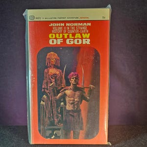 May include: Vintage paperback book titled "Outlaw of Gor" by John Norman. The cover is predominantly orange with illustrations of two figures. The book is sealed in plastic and features the text "Volume II in the Strange History of Counter-Earth."