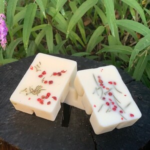 May include: Two white wax melts with a floral design and red berries. The wax melts are shaped like squares with rounded corners.