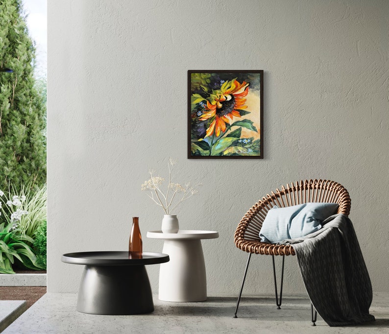 以下が含まれることがあります： オレンジと黄色の花びら、緑の葉、濃い茶色の芯を持つひまわりの絵。ひまわりは濃い茶色の額縁に入っています。