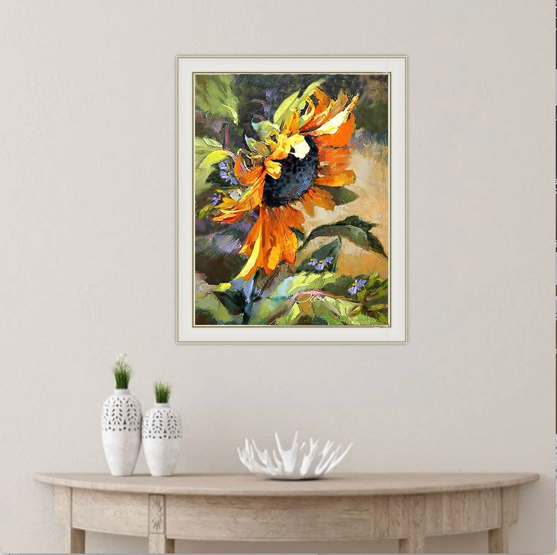 以下が含まれることがあります： 黄色とオレンジの花びら、暗い中心、緑の葉を持つひまわりを描いた額縁付きの絵画。ひまわりは横に傾いており、背景は緑、紫、茶色のぼやけた混合色です。