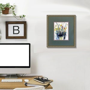 May include: A framed watercolor painting featuring a bouquet of yellow and red wildflowers in a vase. The artwork is encased in a dark green mat with a thin gold frame, displaying a vibrant floral arrangement against a white background.