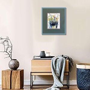 May include: A framed watercolor painting featuring a bouquet of yellow and blue flowers in a vase. The artwork is encased in a dark green mat with a thin red border and a light blue frame, displayed on a wall above a wooden console table.