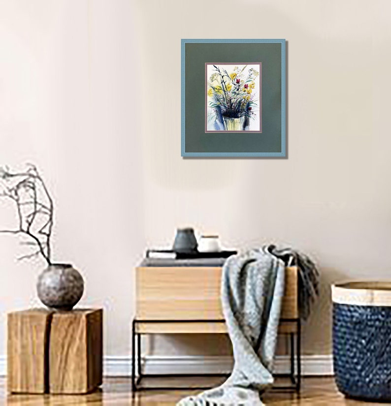 May include: A framed watercolor painting featuring a bouquet of yellow and blue flowers in a vase. The artwork is encased in a dark green mat with a thin red border and a light blue frame, displayed on a wall above a wooden console table.