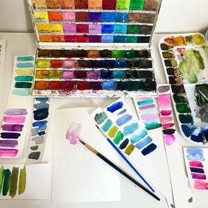 May include: A metal watercolor paint set with multiple rectangular pans of various colors, accompanied by several paper strips featuring hand-painted color swatches, two paintbrushes, and a white sheet of paper with a pink paint stroke.