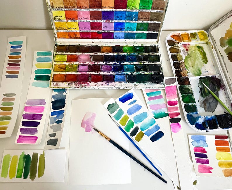 May include: A metal watercolor paint set with multiple rectangular pans of various colors, accompanied by several paper strips featuring hand-painted color swatches, two paintbrushes, and a white sheet of paper with a pink paint stroke.