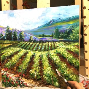 以下が含まれることがあります： ブドウ園の風景を描いた油絵。緑色のブドウの木の列、茶色の土、青い空の下のラベンダーと緑の色合いの丘の背景が特徴です。赤い花が前景にあります。