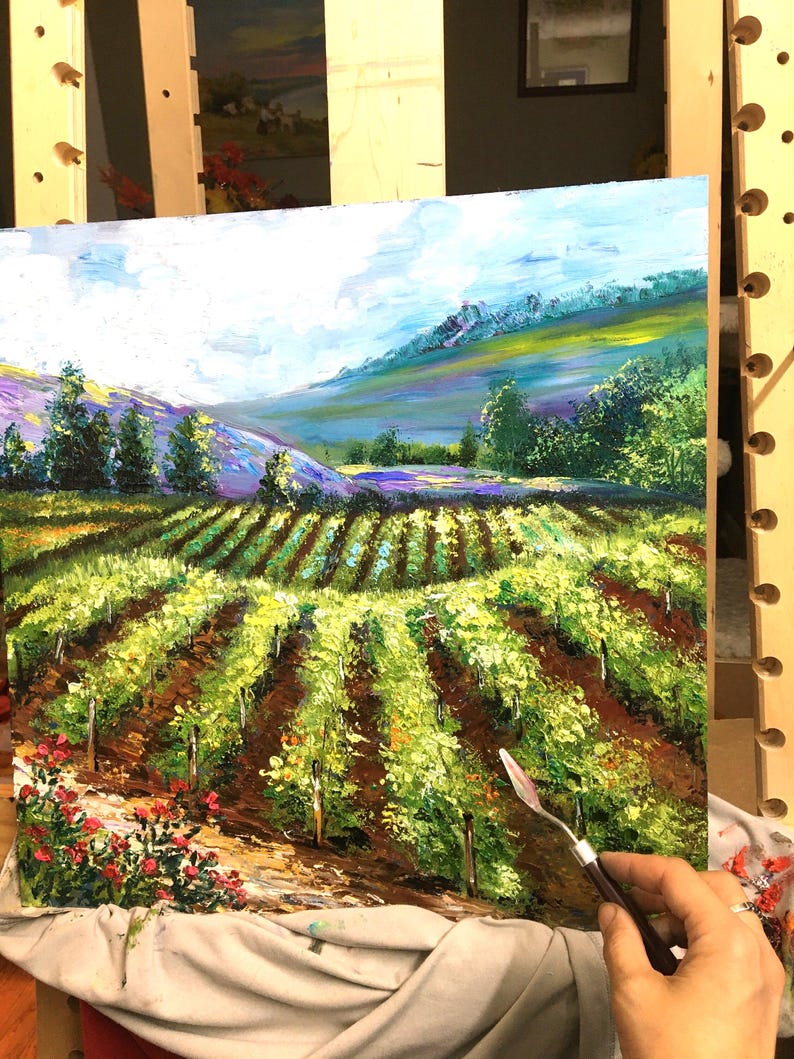 以下が含まれることがあります： ブドウ園の風景を描いた油絵。緑色のブドウの木の列、茶色の土、青い空の下のラベンダーと緑の色合いの丘の背景が特徴です。赤い花が前景にあります。