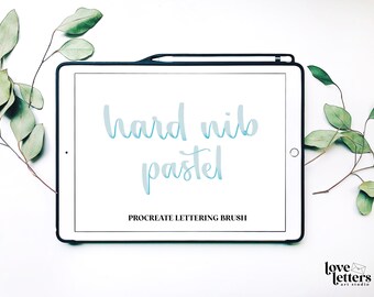 May include: A white tablet screen with the text "hard nib pastel" in blue and the text "PROCREATE LETTERING BRUSH" in black. The tablet is on a white surface with green leaves on either side.