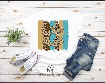 Puede incluir: Camiseta blanca con un diseño gráfico de las palabras "RANCH HAND" en letras doradas sobre un fondo a rayas azul y marrón con detalles de estampado de leopardo.