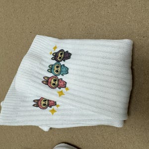 May include: White ribbed knit socks with cartoon bunny characters in purple, blue, pink, and yellow, with star accents. The socks are folded.