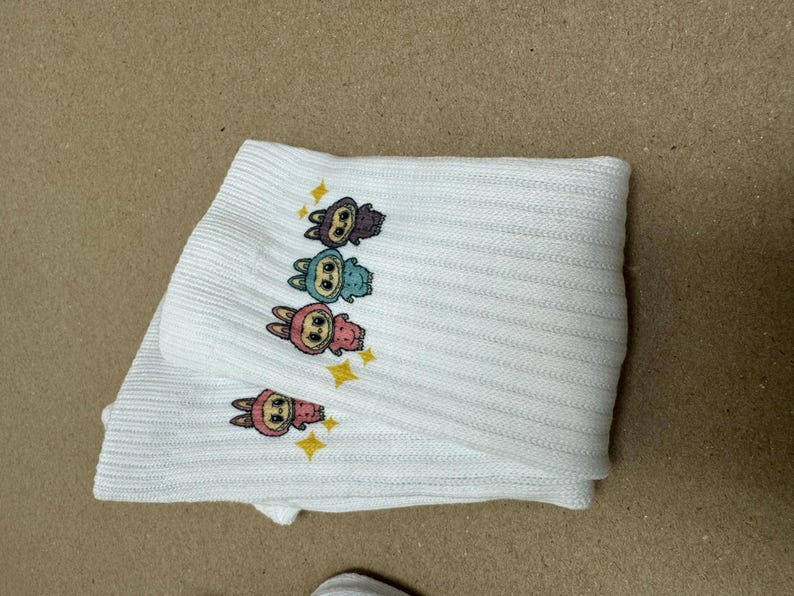 May include: White ribbed knit socks with cartoon bunny characters in purple, blue, pink, and yellow, with star accents. The socks are folded.