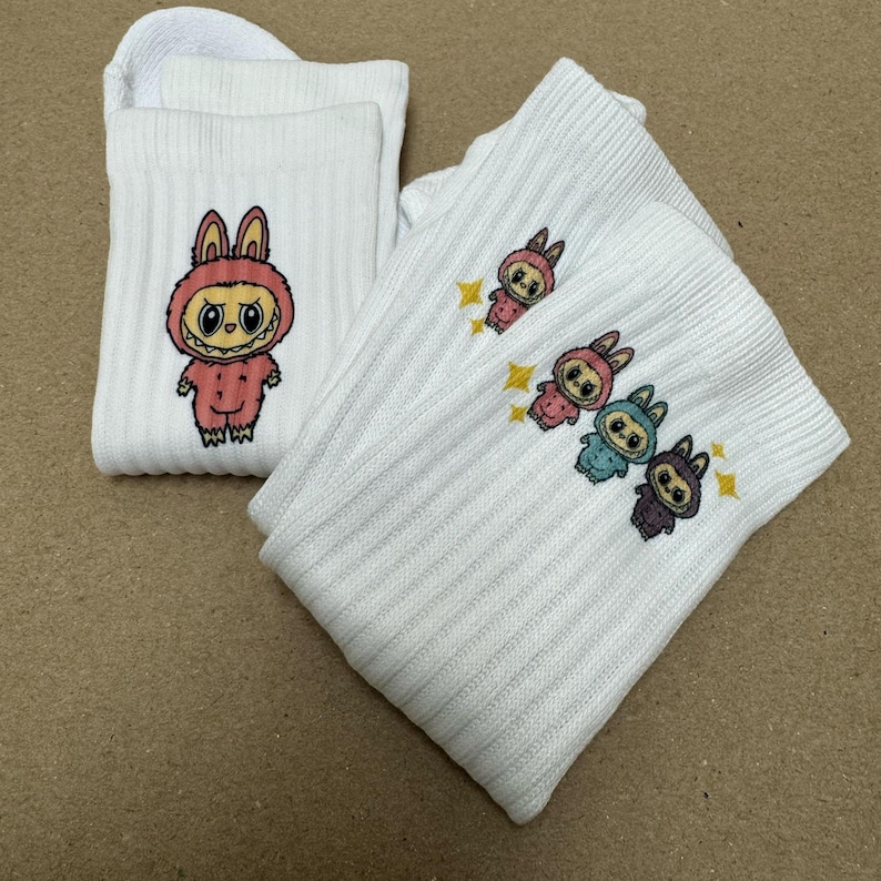 May include: White ribbed socks with cartoon bunny characters in pink, blue, and purple outfits and yellow star accents. One sock shows a single bunny, the other has three.