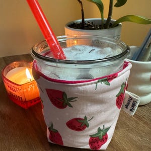 May include: A clear glass with a red straw, filled with a white beverage and ice. The glass is wrapped in a pink fabric cozy with a strawberry pattern. A lit candle and potted plants are in the background.