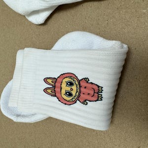 May include: White socks featuring a cartoon character. The character is a pink, furry creature with a yellow face, large eyes, and bunny ears. The socks are folded and placed on a light brown surface.