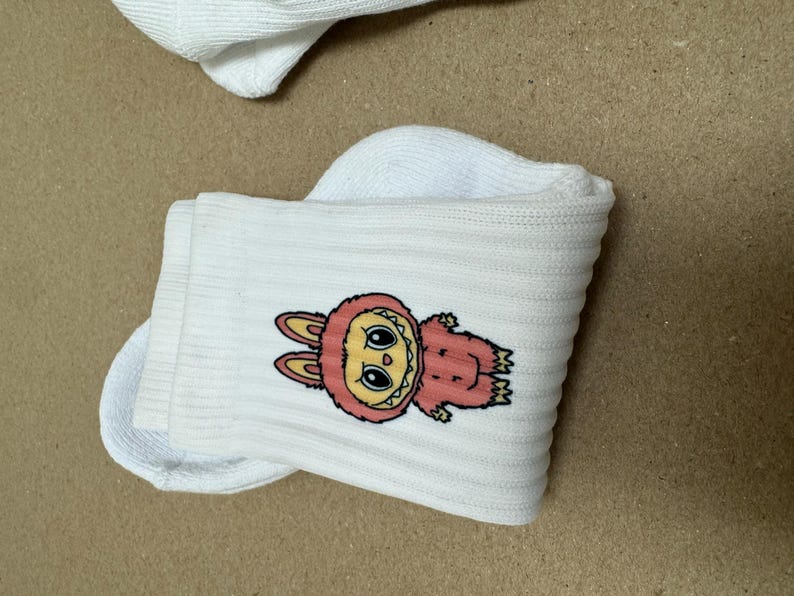 May include: White socks featuring a cartoon character. The character is a pink, furry creature with a yellow face, large eyes, and bunny ears. The socks are folded and placed on a light brown surface.