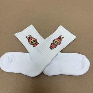 May include: Two pairs of white crew socks featuring a cartoon bunny design. The bunny is dressed in a pink hooded outfit and has large eyes. The socks are made of a soft, ribbed material.