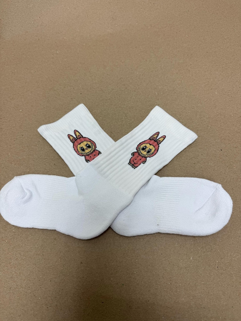 May include: Two pairs of white crew socks featuring a cartoon bunny design. The bunny is dressed in a pink hooded outfit and has large eyes. The socks are made of a soft, ribbed material.