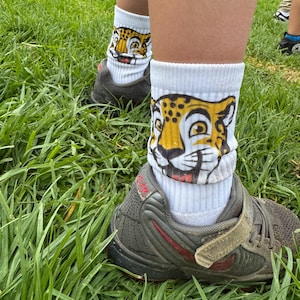 May include: White athletic socks featuring a cartoon jaguar face. The socks are paired with grey and red trainers, set against a background of green grass.