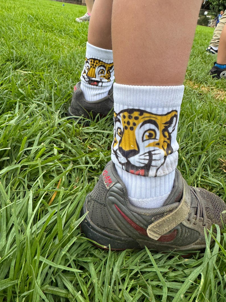 May include: White athletic socks featuring a cartoon jaguar face. The socks are paired with grey and red trainers, set against a background of green grass.