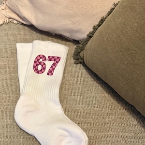 May include: White athletic socks with the number 67 in a pink and purple checkered pattern. The socks are folded and resting on a gray textured surface, with pillows in the background.