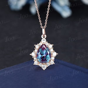 May include: Rose gold pendant necklace featuring a pear-shaped, color-changing alexandrite gemstone, surrounded by a halo of small, round, white gemstones. The necklace has a delicate chain and a secure clasp. The pendant is approximately 1 inch in length.