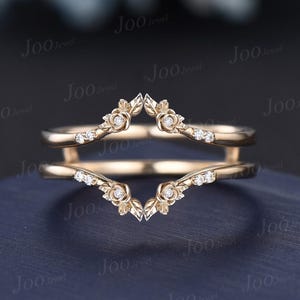 Może przedstawiać: Złota osłona na pierścionek z kwiatowym wzorem. Pierścionek posiada małe, błyszczące kamienie szlachetne i akcenty w kształcie róży. Opaska zakrzywia się wokół głównego pierścionka, tworząc niepowtarzalny i elegancki wygląd. Tło jest ciemnoniebieskie.