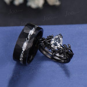 Puede incluir: Un juego de anillos de boda negro con un anillo de compromiso de diamante de forma pera. El anillo de compromiso presenta una banda de metal negro con un delicado diseño de hojas y detalles negros.