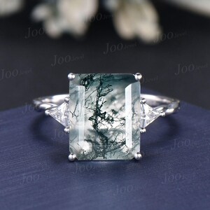 May include: A silver ring with a large, rectangular moss agate gemstone, displaying a distinctive pattern of green and white inclusions. Two smaller, triangular clear gemstones are set either side of the main stone. The ring band has a twisted design.