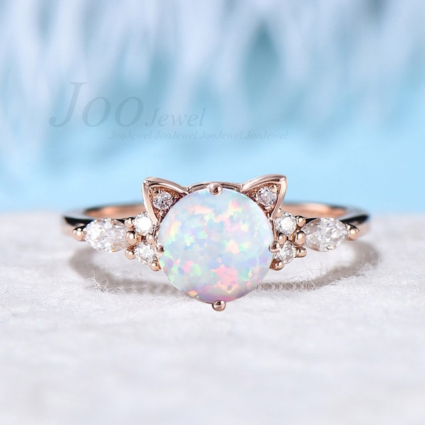 Op de afbeelding: Een ring van roségoud met een witte opaal als middelste steen en kleine heldere stenen rond de band. De ring heeft een kattenoor-ontwerp met een kleine heldere steen in elk oor.