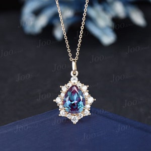 May include: A gold-colored pendant necklace featuring a pear-shaped, color-changing gemstone, surrounded by small, clear gemstones. The pendant hangs from a delicate gold chain. The background is dark blue.