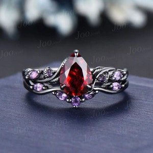 Op de afbeelding: Een zwarte ringenset met een peervormige, dieprode edelsteen als middelpunt. De ring is geaccentueerd met kleinere, ovaalvormige paarse edelstenen en een bladachtig ontwerp. De ring staat tegen een donkerblauwe achtergrond.