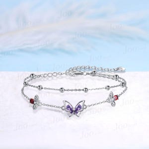 May include: A silver double-strand bracelet featuring a butterfly and floral design. The butterfly has purple gemstones, and the flowers have red gemstones. The bracelet has a delicate chain with small silver beads.