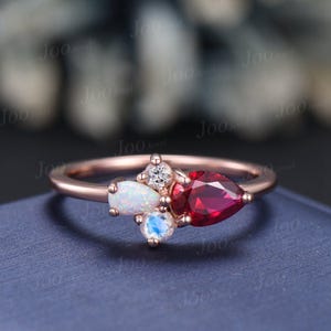 May include: A rose gold ring with a cluster of gemstones. The ring features a pear-shaped red gemstone, a white opal, a round white gemstone, and a moonstone.