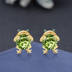 May include: A pair of gold-coloured frog-shaped earrings. Each earring features a large, round, light green gemstone for the body, with small blue gemstones for the eyes. The frog's legs and head are crafted in gold.