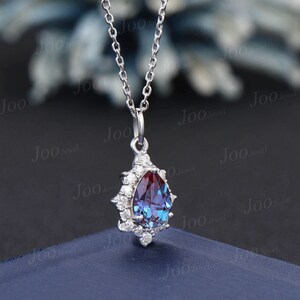 May include: A silver necklace with a pear-shaped pendant. The pendant features a color-changing gemstone, transitioning from purple to blue, surrounded by small, clear gemstones. The necklace is displayed on a dark blue surface.