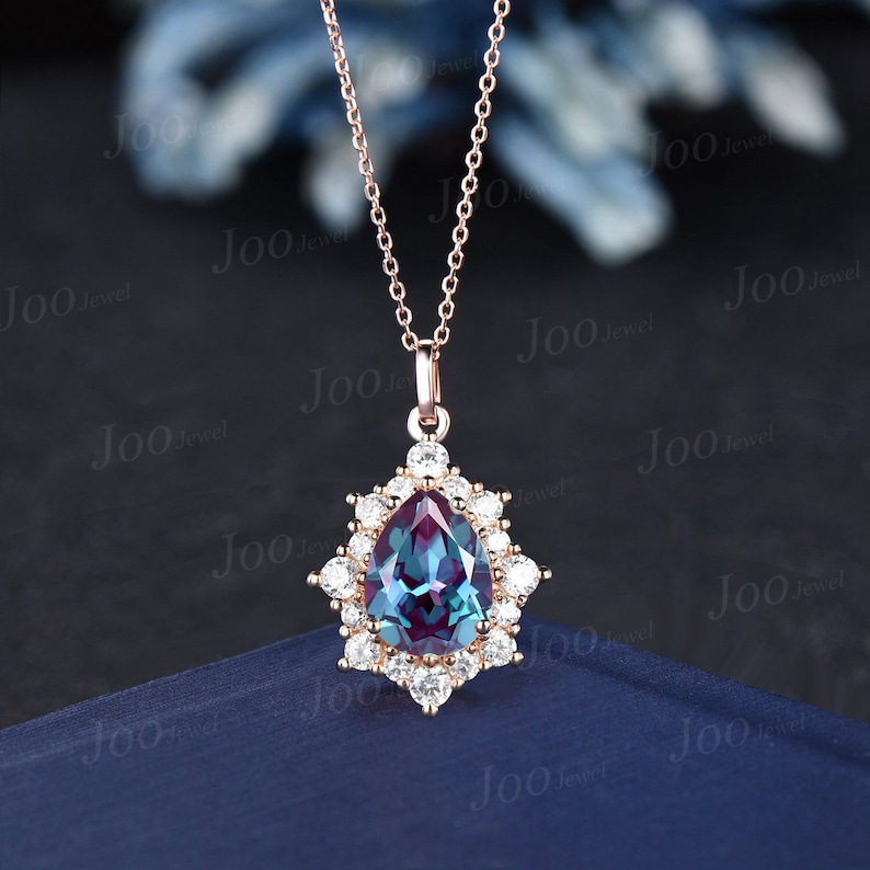 May include: Rose gold pendant necklace featuring a pear-shaped, color-changing alexandrite gemstone, surrounded by a halo of small, round, white gemstones. The necklace has a delicate chain and a secure clasp. The pendant is approximately 1 inch in length.