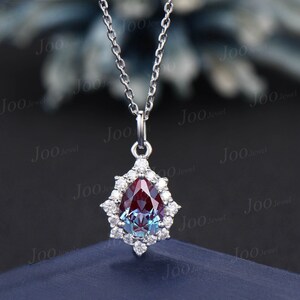 May include: A silver necklace featuring a pear-shaped pendant with a color-changing alexandrite gemstone. The gemstone is surrounded by a halo of small, round diamonds. The chain is delicate and the pendant hangs from a small bail.