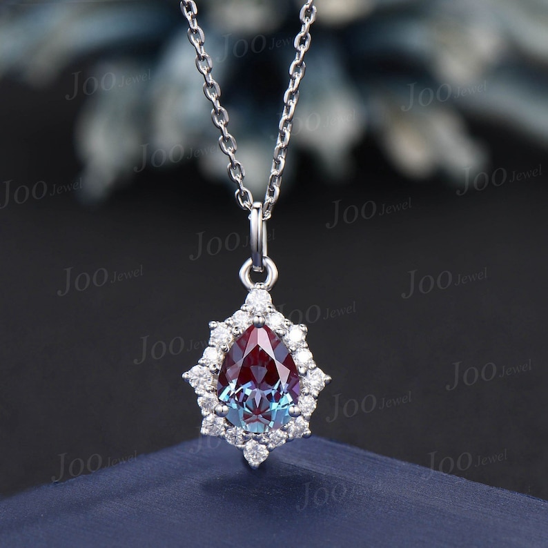 May include: A silver necklace featuring a pear-shaped pendant with a color-changing alexandrite gemstone. The gemstone is surrounded by a halo of small, round diamonds. The chain is delicate and the pendant hangs from a small bail.