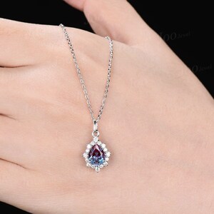May include: A silver necklace with a pear-shaped pendant. The pendant features a color-changing gemstone, surrounded by small, clear gemstones. The chain is delicate and the pendant is held by a small bail.