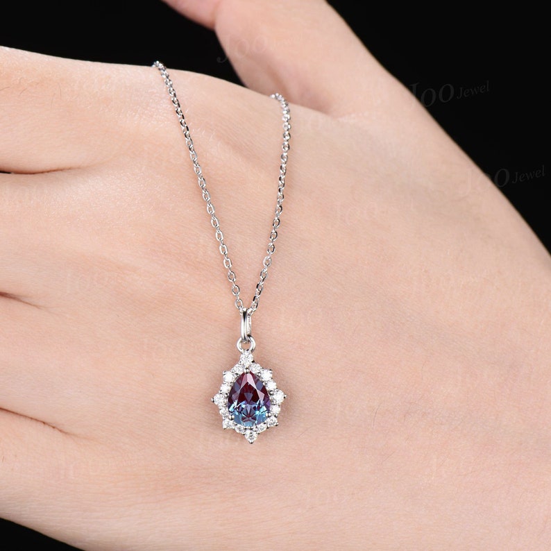 May include: A silver necklace with a pear-shaped pendant. The pendant features a color-changing gemstone, surrounded by small, clear gemstones. The chain is delicate and the pendant is held by a small bail.