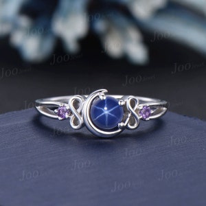 May include: A silver ring featuring a large, round, blue star sapphire gemstone. The ring has a crescent moon design with two smaller purple gemstones on either side. The ring is set against a dark blue background.