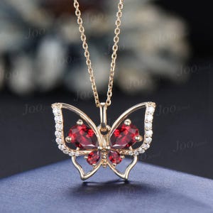 Op de afbeelding: Een gouden vlinderketting met rode edelsteenaccenten. De vlindervleugels zijn versierd met kleine, heldere stenen.